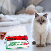 On a wooden floor, a fluffy white and grey cat is captivated by a hand flipping through the pages of the "50 Ways Your CAT is Trying to KILL YOU! Funny Desk Spiral Flip Book Signs," making it an ideal gift for cat lovers who are fascinated by all things feline.