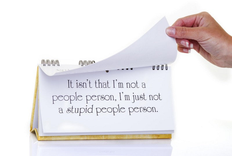 Hand flipping a page of the CLASSIC INSULTS Desk Spiral Flip Book - Funny Office Message Table Signs with text: "It isn't that I'm not a people person, I'm just not a stupid people person." This humorous novelty gift is perfect for those who appreciate professional insults with a dash of wit.