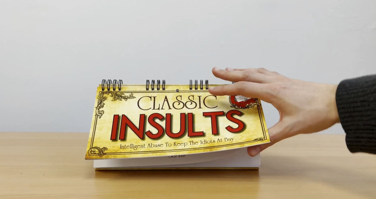 A hand flipping a page of the "CLASSIC INSULTS Desk Spiral Flip Book - Funny Office Message Table Signs" rests on a wooden table, making it the perfect novelty gift for those who appreciate witty banter.