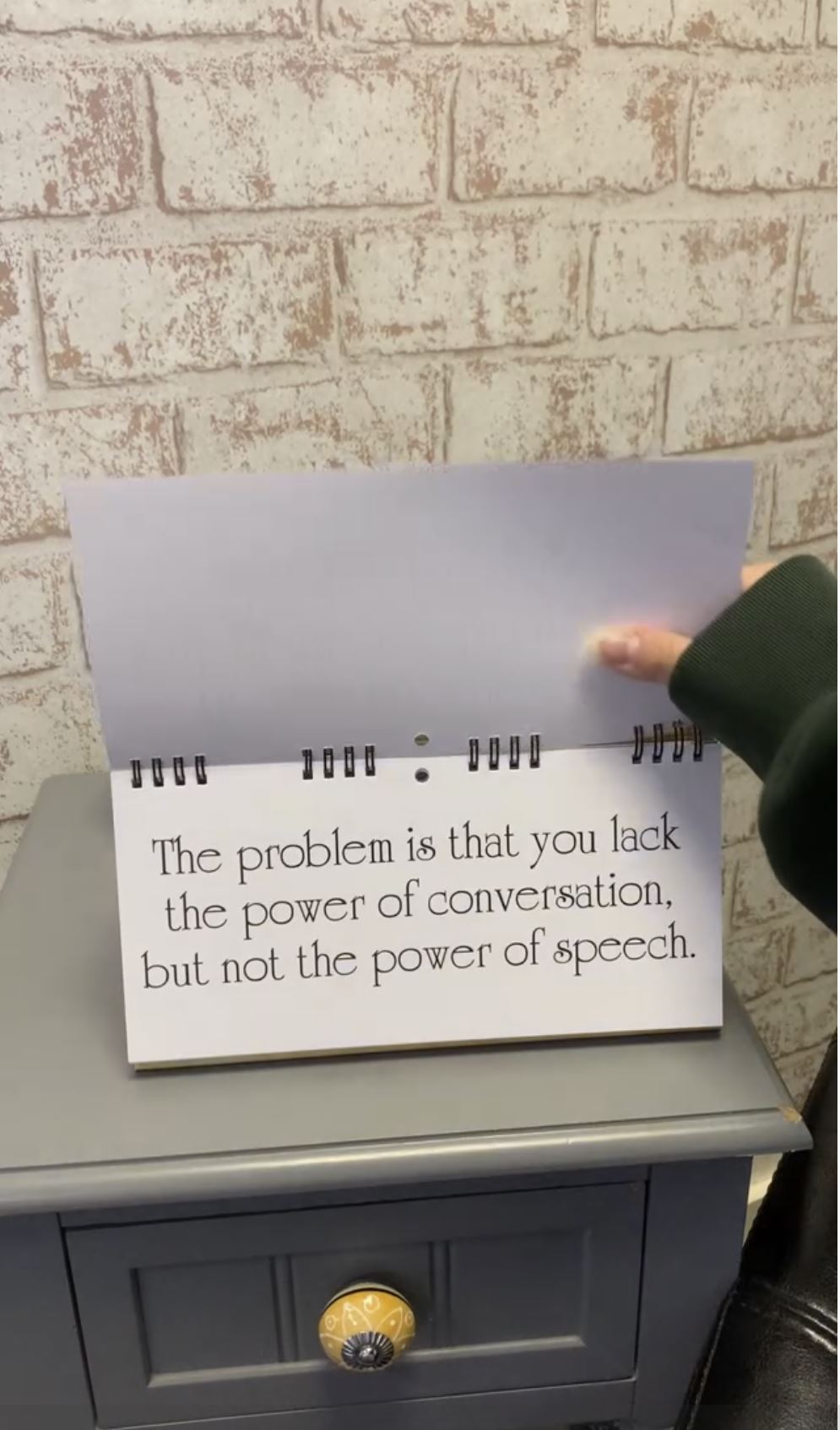 Close-up of a hand flipping a CLASSIC INSULTS Desk Spiral Flip Book with the message: "The problem is that you lack the power of conversation, but not the power of speech." This humorous office message table sign makes for a perfect novelty gift to bring laughter to any desk.