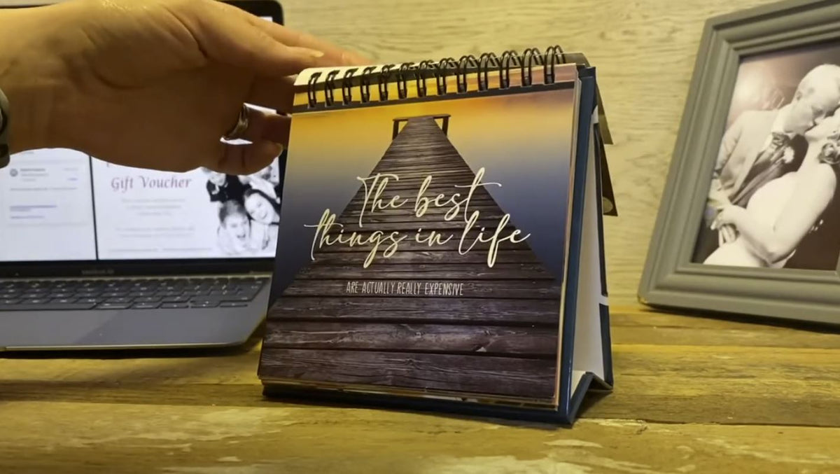 A small Demotivational Quotes Desk Spiral Flip Book - Hysterical Message Table Signs, showcasing quotes like "The best things in life are actually really expensive," sits next to a laptop and a framed photo. This quirky novelty gift adds a touch of humor to your workspace, making it uniquely personal.