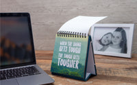 A desk features a laptop and a Demotivational Quotes Desk Spiral Flip Book, humorously proclaiming "When the going gets tough, the tough get tougher," making it an ideal novelty gift. Nearby, a framed photo of two smiling people adds warmth to the scene.