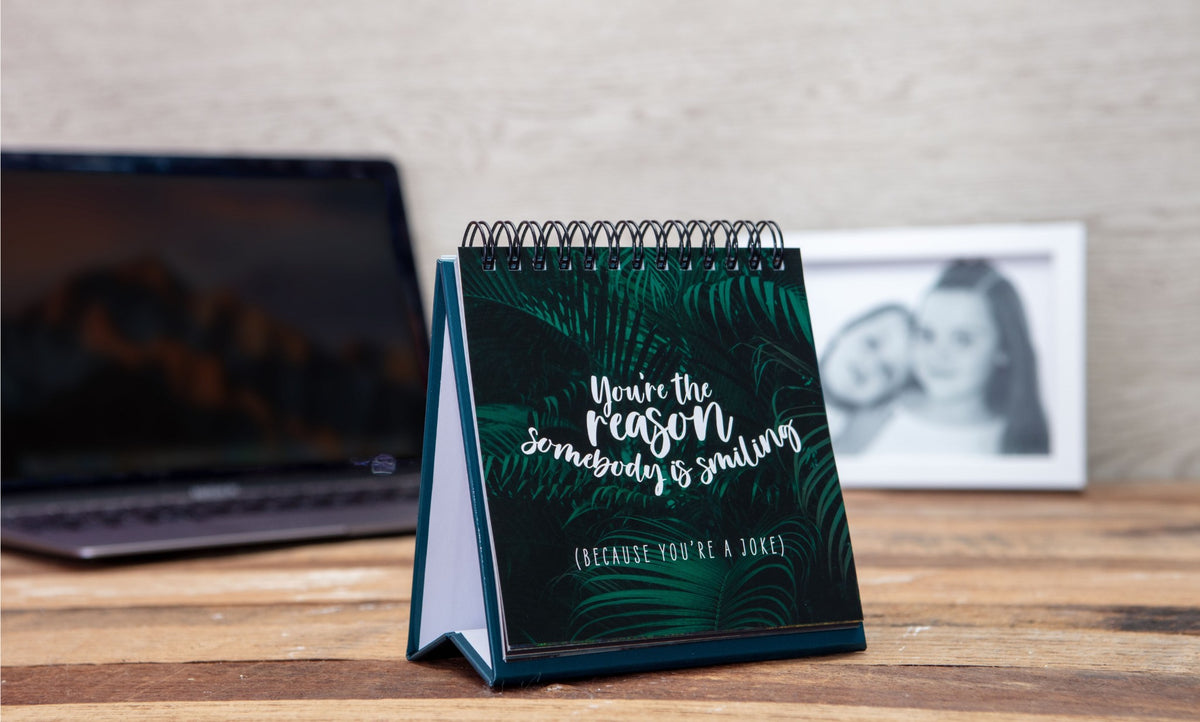 A desk displays a spiral calendar with a motivational quote, while in the background, a blurred laptop and a monochromatic photo of two people can be seen. Beside it sits the Demotivational Quotes Desk Spiral Flip Book - Hysterical Message Table Signs, providing cheeky novelty gifts of demotivational quotes for those light-hearted moments.