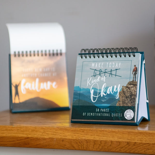 Two spiral-bound books rest on a wooden surface. The left book, titled "Demotivational Quotes Desk Spiral Flip Book - Hysterical Message Table Signs," features a blurred outdoor image with the word "failure," embracing demotivational quotes. The right book, perfect as a novelty gift or desktop flipbook, says "Make Today Kind of Okay" with a mountain climber graphic.