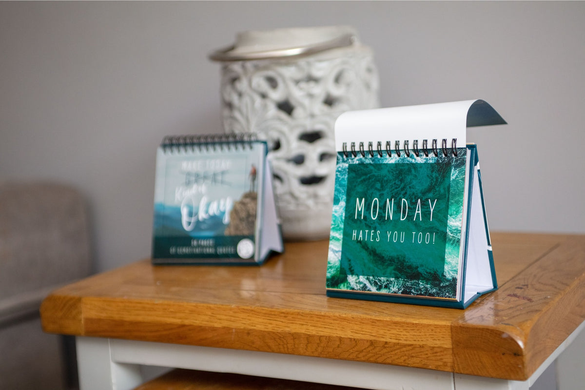 Two desk calendars on a wooden table, one featuring the "Demotivational Quotes Desk Spiral Flip Book - Hysterical Message Table Signs" with the quote "Monday hates you too!" and the other showcasing an abstract landscape, make for a perfect novelty gift or desktop flipbook that adds personality to any workspace.