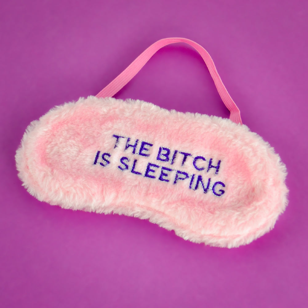 The pink "THE BITCH IS SLEEPING" mask, featuring blue embroidered text, sits on a purple background. It's the perfect funny sleep accessory and a delightful novelty gift.