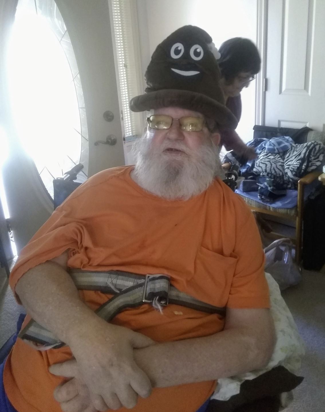An older man with a white beard and glasses wears an orange shirt and the 💩 Poop Emoji Hat – Funny Gag Gift & Hilarious Party Costume Accessory, while sitting indoors with a woman in the background.