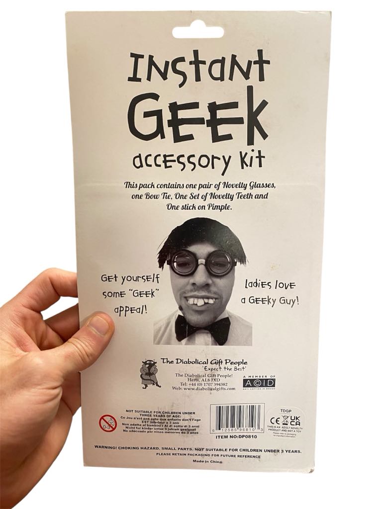 A hand holds the Instant Geek Costume Kit package, featuring illustrations of glasses, fake teeth, bow tie, and pimple—funny gag gifts and perfect accessories for a nerd dress up set—with witty descriptions and novelty warnings.