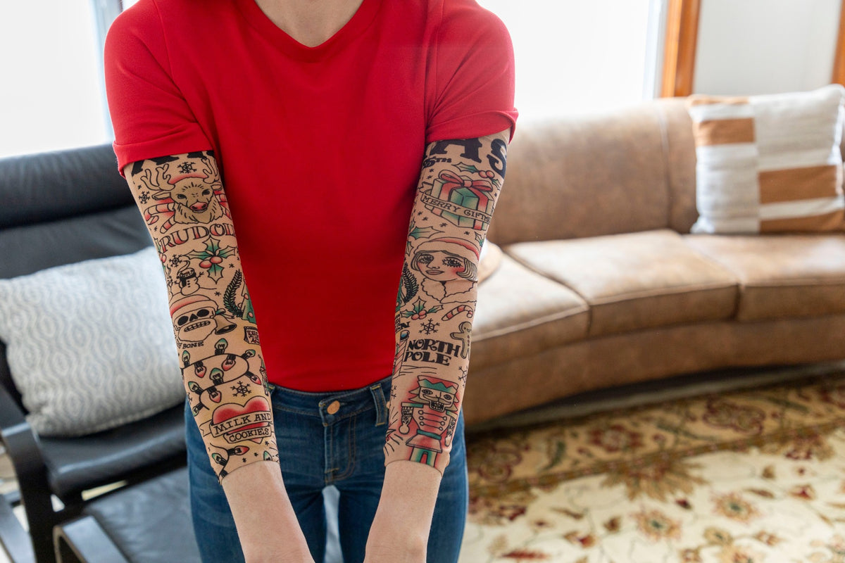 Relaxing on a leather couch, a person in a red t-shirt shows off both arms covered with the 🎄 Christmas Holiday Tattoo Sleeves 2pk – Funny Fake Arm Tattoos Costume Party, adding festive flair to their holiday look.