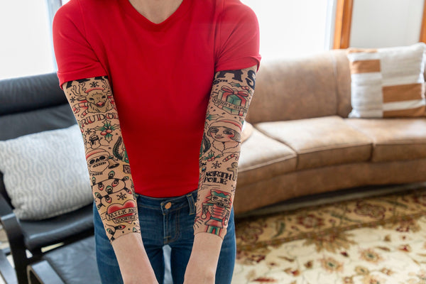 Relaxing on a leather couch, a person in a red t-shirt shows off both arms covered with the 🎄 Christmas Holiday Tattoo Sleeves 2pk – Funny Fake Arm Tattoos Costume Party, adding festive flair to their holiday look.