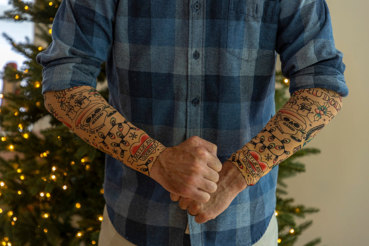 A person poses in front of a Christmas tree wearing 🎄 Christmas Holiday Tattoo Sleeves 2pk—funny fake arm tattoos that make a perfect festive costume accessory for any holiday party.