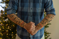 A person poses in front of a Christmas tree wearing 🎄 Christmas Holiday Tattoo Sleeves 2pk—funny fake arm tattoos that make a perfect festive costume accessory for any holiday party.