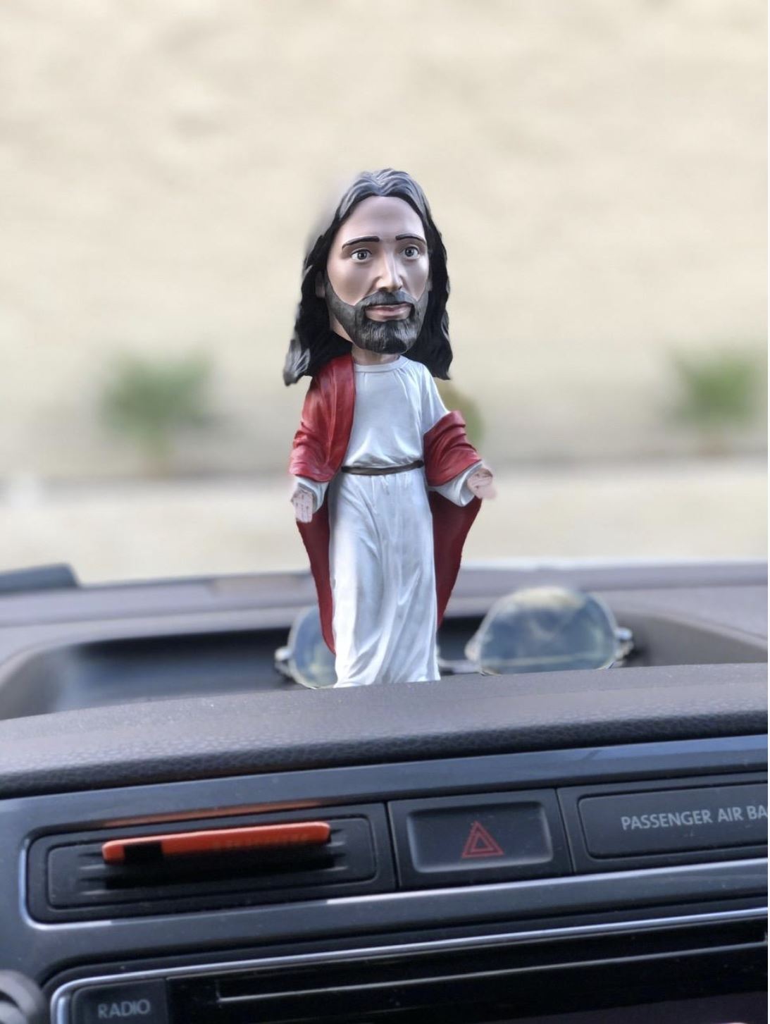 The 🙏 Jesus Bobblehead Figure – Premium Lifelike Collectible Gift by Royal Bobbles✝️ rests on the car dashboard, with control buttons and a radio below.