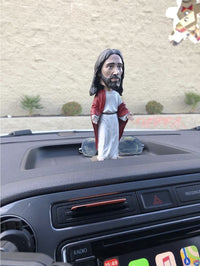 The 🙏 Jesus Bobblehead Figure – Premium Lifelike Collectible Gift by Royal Bobbles✝️ features a hand-painted Jesus in a red and white robe sitting on a dashboard, with sunglasses behind it and a beige wall in the background.