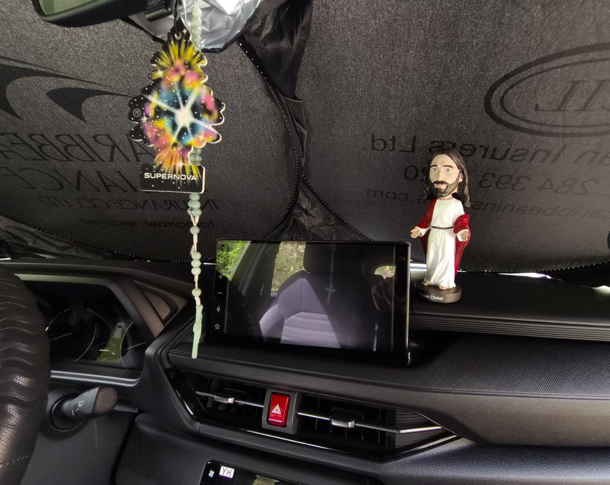 A car dashboard displays a dangling air freshener, a sunshade on the windshield, and a hand-painted 🙏 Jesus Bobblehead Figure – Premium Lifelike Collectible Gift | Royal Bobbles✝️.