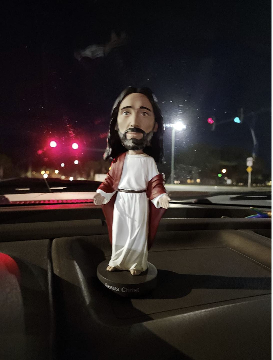 A 🙏 Jesus Bobblehead Figure – Premium Lifelike Collectible Gift by Royal Bobbles✝️ stands on a car dashboard at night as red traffic lights glow through the windshield.