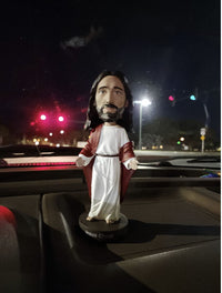 A 🙏 Jesus Bobblehead Figure – Premium Lifelike Collectible Gift by Royal Bobbles✝️ stands on a car dashboard at night as red traffic lights glow through the windshield.