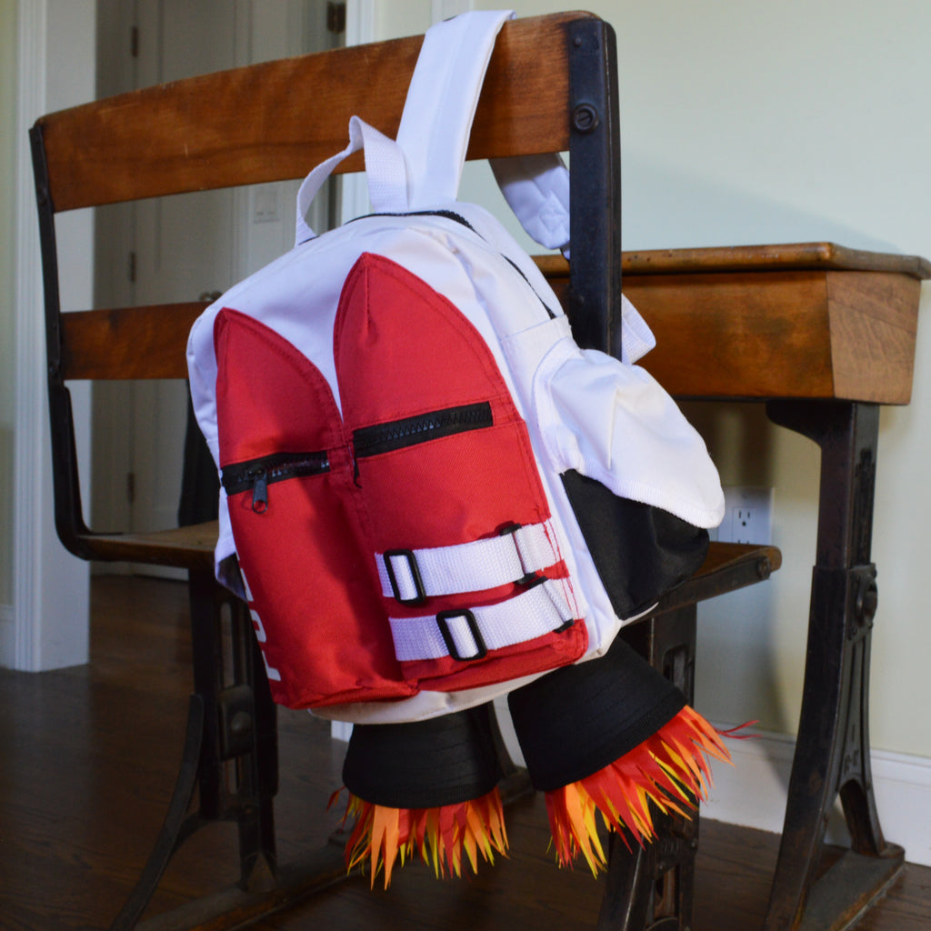 The Kids Jetpack Backpack Rocket School Bag 🚀, featuring red fins and flame details, hangs on the back of a wooden chair—perfect for space-loving kids or as an astronaut costume accessory and travel gift.