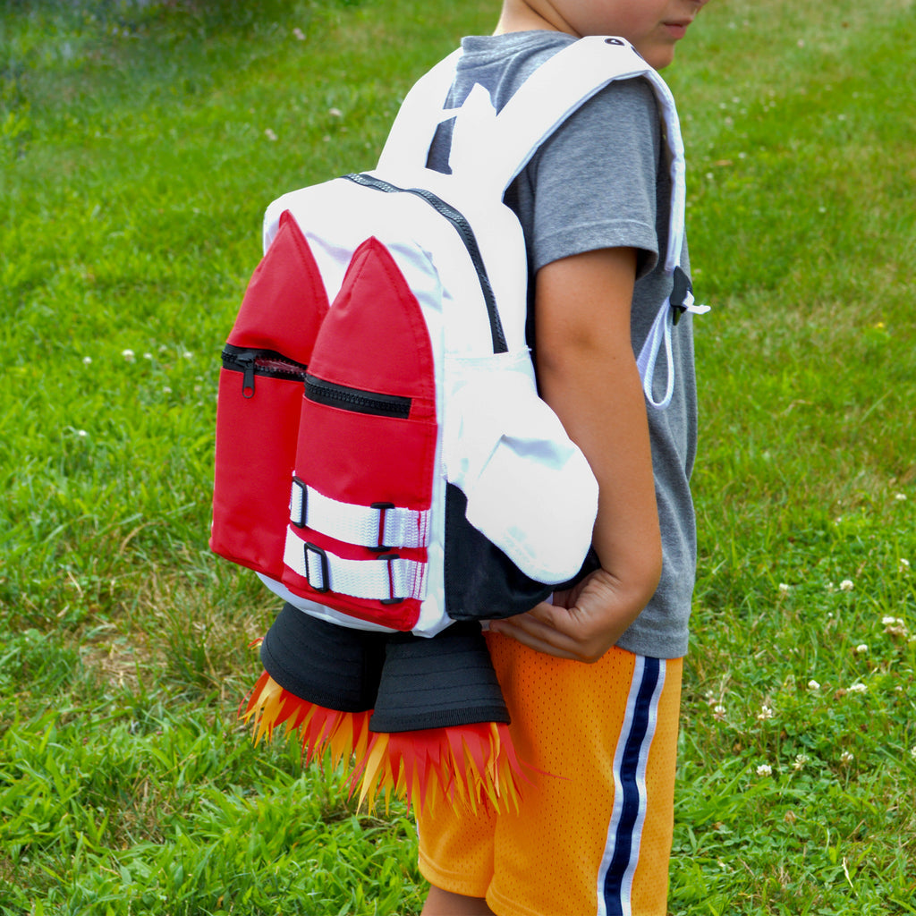 A child stands on grass wearing a grey shirt, yellow athletic shorts, and the Kids Jetpack Backpack Rocket School Bag 🚀—a space astronaut costume travel gift with flame details.