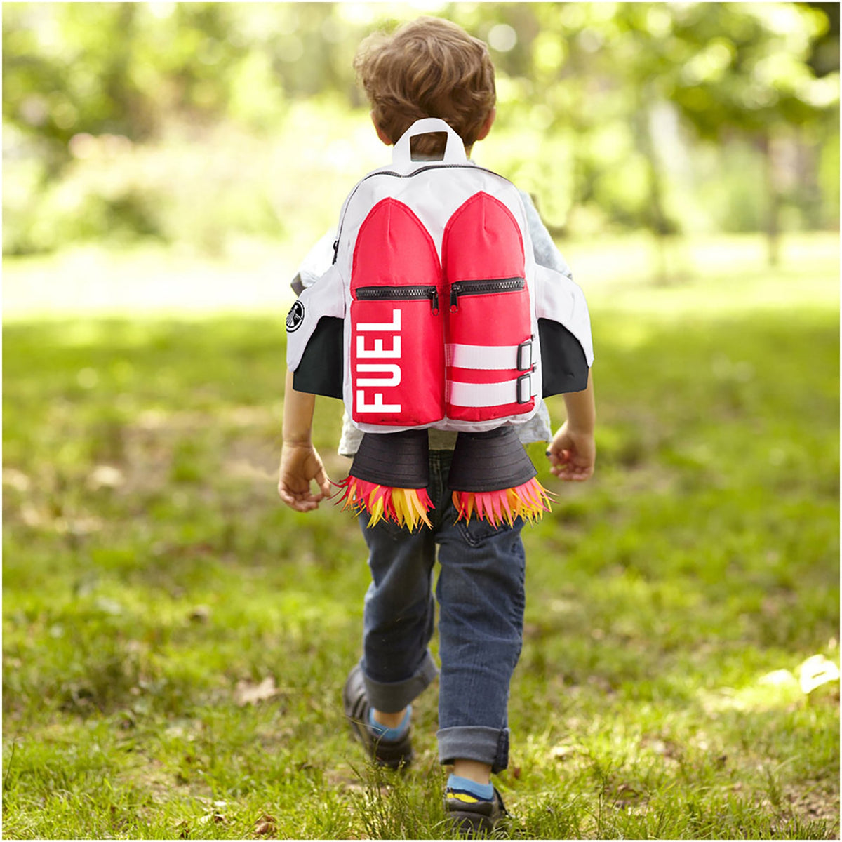 A child walks on grass wearing jeans, a white jacket, and the Kids Jetpack Backpack Rocket School Bag 🚀, designed to look like a jetpack with "FUEL" text and fabric flames, perfect for space-themed adventures.