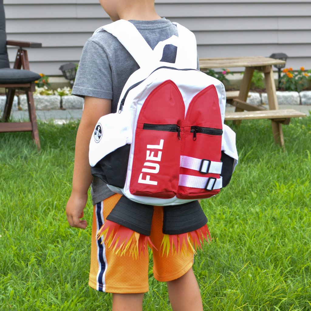 A child stands on grass in a backyard wearing the Kids Jetpack Backpack Rocket School Bag 🚀, designed like a jetpack with "FUEL" and flame details—perfect for space astronaut costumes or as a fun travel gift.
