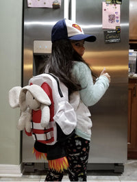 A young girl in a Superman cap and patterned leggings stands in a kitchen with the Kids Jetpack Backpack Rocket School Bag, holding a stuffed bunny and giving a thumbs up.