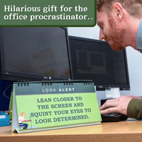 As a man leans toward the computer screen with determination, a desk calendar displays the message: "Lean closer to the screen and squint your eyes to look determined." Perhaps he's browsing through humorous suggestions from the "How to 'LOOK BUSY' at Work - Slacker's Guide - Funny Desk Spiral Flip Book Signs" for a perfect Secret Santa gift.