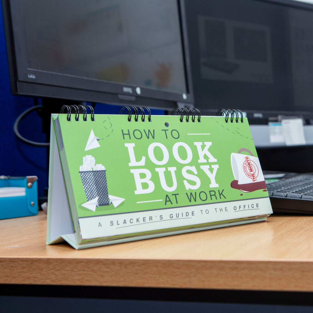 A desk accessory, the "How to 'LOOK BUSY' at Work - Slacker's Guide - Funny Desk Spiral Flip Book Signs," sits on an office desk amidst computers in the background. This humorous novelty is perfect as a funny office gift or a Secret Santa surprise, filled with entertaining ideas to keep colleagues amused and guessing.