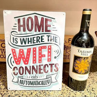 A bottle of wine is paired with the charming Metal Tin Sign "Home is Where the WiFi Connects Automatically" Wall Decor #H-101, adding a whimsical touch to your home decor and serving as a clever gift idea for friends.