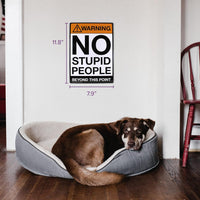 A dog rests in a gray pet bed on a wooden floor. Above, the Metal Tin Sign "Warning: NO STUPID PEOPLE Beyond This Point" Bar Man Cave #C-101 hangs humorously, measuring 11.8 inches tall and 7.9 inches wide.