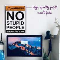 On the wall, there is a Metal Tin Sign labeled "Warning: NO STUPID PEOPLE Beyond This Point" (#C-101), which adds a hint of novelty decor to the room. Below it, a desk is adorned with a monitor, lamp, Eiffel Tower model, and cactus. The description guarantees that this playful sign's high-quality print will remain vibrant over time.