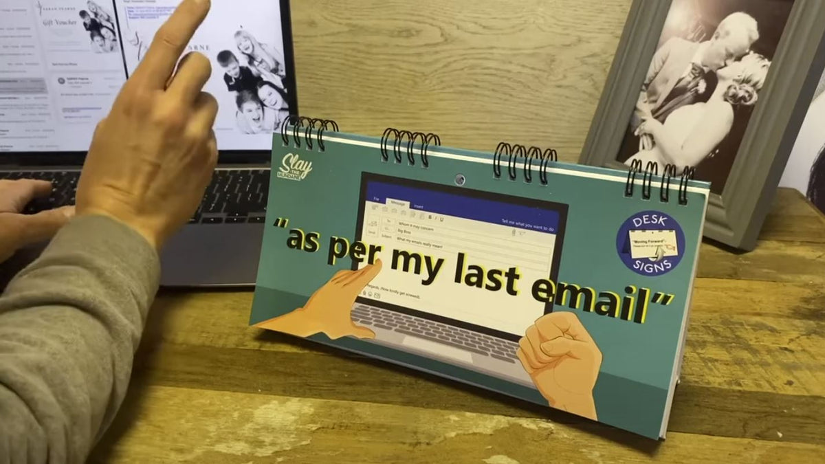 A person gestures at a laptop, next to a desk sign from the "As Per My Last Email - Desk Spiral Flip Book - Funny Office Message Table Signs." This novelty flip book offers sarcastic translations that enhance the office humor, complemented by framed photos in the background.