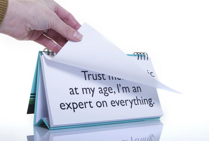 A hand flips through the pages of the Senior Moments Desk Spiral Flip Book, showcasing the phrase, "Trust me, at my age, I'm an expert on everything," making it a perfect funny retirement gift or white elephant surprise.