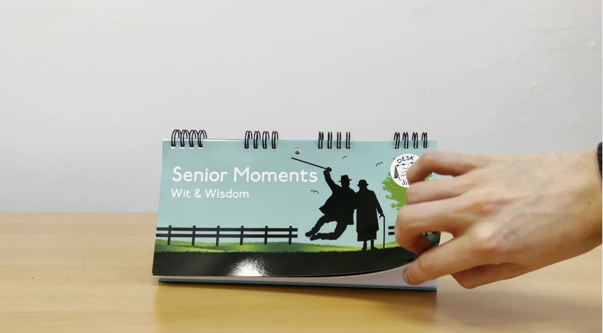 A hand flips through a desk calendar called "Senior Moments - Desk Spiral Flip Book – Over Hill Message Table Desk Signs," showcasing a silhouetted golfing scene, making it an ideal humorous retirement gift or a perfect addition to your collection of white elephant gifts.