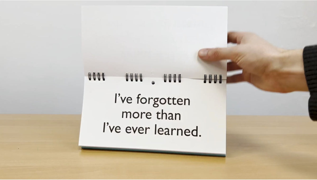 A hand holds a spiral-bound flip chart from the "Senior Moments - Desk Spiral Flip Book – Over Hill Message Table Desk Signs," displaying the text, "I've forgotten more than I've ever learned," on a wooden table. It's perfect as a funny retirement gift or a clever addition to your collection of white elephant gifts.