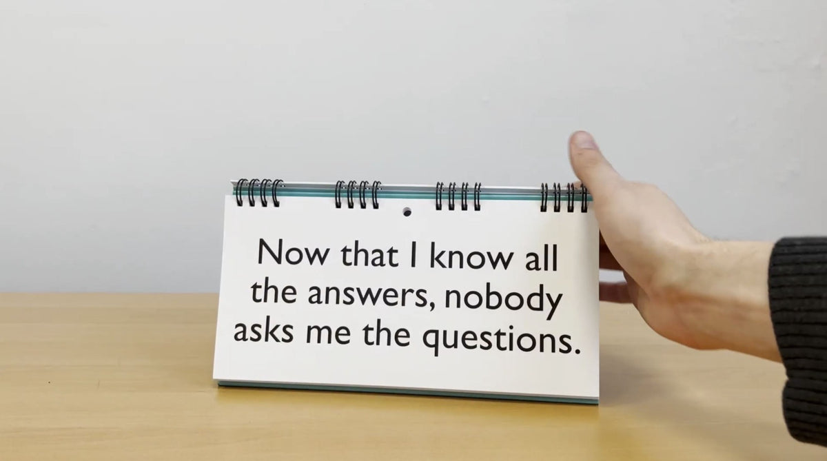 A hand holds the "Senior Moments - Desk Spiral Flip Book – Over Hill Message Table Desk Signs," showcasing the text: "Now that I know all the answers, nobody asks me the questions." This makes for a perfect humorous retirement gift, combining wit and wisdom for those embarking on their next adventure.