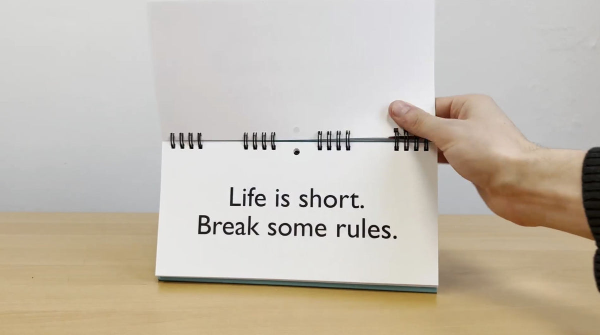 A person holds a spiral-bound flipbook, the "Senior Moments - Desk Spiral Flip Book – Over Hill Message Table Desk Signs," open to a page that reads, "Life is short. Break some rules." It's an ideal choice for a funny retirement gift or the next white elephant gift exchange, offering a lighthearted reminder to inject fun into life.