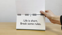 A person holds a spiral-bound flipbook, the "Senior Moments - Desk Spiral Flip Book – Over Hill Message Table Desk Signs," open to a page that reads, "Life is short. Break some rules." It's an ideal choice for a funny retirement gift or the next white elephant gift exchange, offering a lighthearted reminder to inject fun into life.