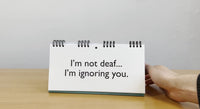 A hand confidently holds a Senior Moments desk spiral flip book on a wooden surface, with a clever message that reads, "I'm not deaf... I'm ignoring you"—an ideal humorous retirement gift for those embracing their golden years.