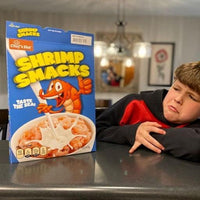 A young person in a red and black sweater leans on a kitchen counter next to a large 🦐 Prank Cereal Box – Shrimp Smacks Gag Gift Box, featuring a cartoon shrimp on the funny joke prank present packaging.