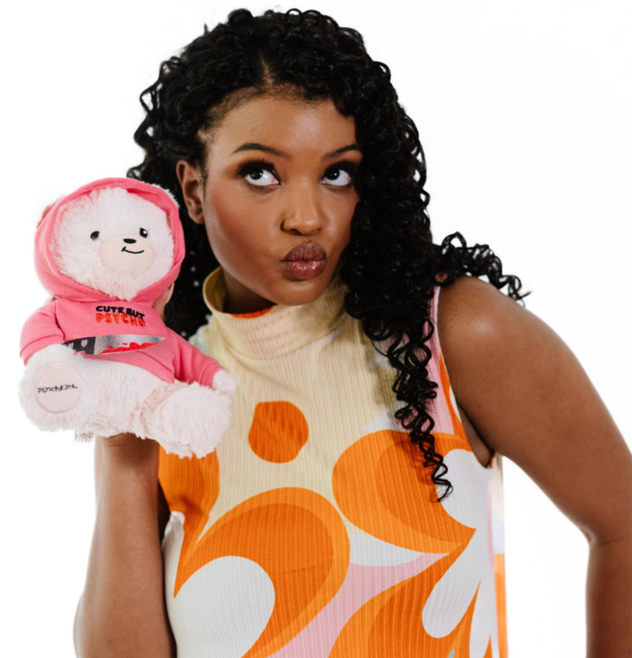 A woman with curly hair holds a Punchkins "Cute But PSYCHO" Teddy Bear Plushie, featuring a pink hoodie and bloody kitchen knife, and smiles playfully while showing off her quirky gift.