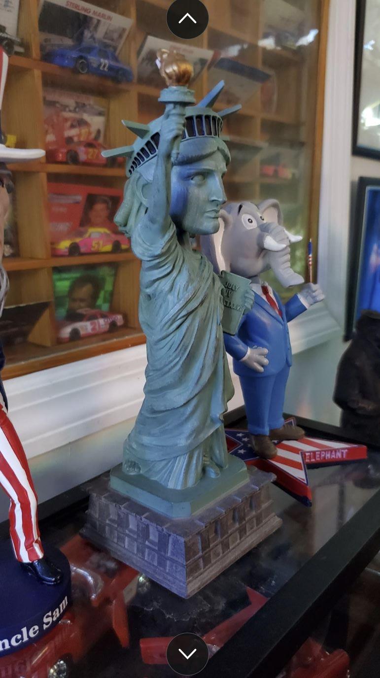A Statue of Liberty Bobblehead Figure 🗽 – Premium Lifelike Collectible Decor Gift sits on a shelf beside a cartoon elephant in a suit, with model cars behind—a playful collectible scene for any enthusiast.