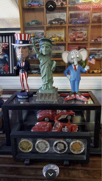 Three figurines—an Uncle Sam, a Republican elephant, and the Statue of Liberty Bobblehead Figure 🗽 – Premium Lifelike Collectible Decor Gift—are displayed before a case of red toy cars and collectible coins, with model cars on a shelf above.