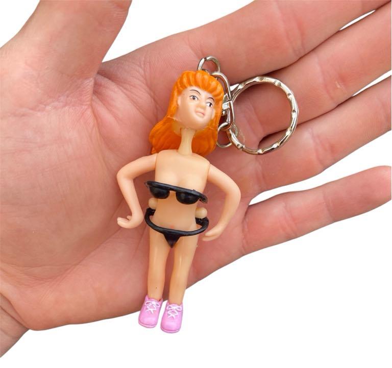 A hand holds the Saucy Suzie Flashing Stripper Keychain—an adult gag gift featuring a small plastic woman with orange hair, black bikini, and pink shoes; perfect for bachelorette parties or cheeky pranks.