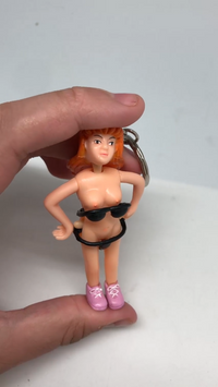 A hand holds the Saucy Suzie Flashing Stripper Keychain, a funny adult gag gift featuring a small figurine of a woman in black underwear and pink shoes with orange hair—perfect as a playful peepshow toy.