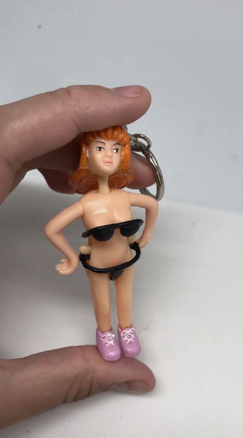 A hand holds the Saucy Suzie Flashing Stripper Keychain—a funny adult gag gift peepshow toy featuring a woman with orange hair, black bikini, and pink shoes—against a plain white background.