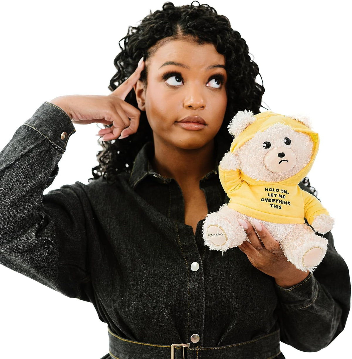 A woman in a black denim shirt points to her head while holding the Punchkins 🐻 "Let Me Overthink This" Teddy Bear Plushie—an ideal gift for overthinkers, featuring a funny yellow hoodie with a witty phrase.