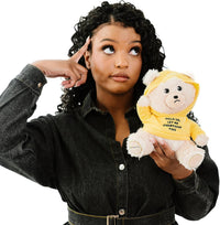 A woman in a black denim shirt points to her head while holding the Punchkins 🐻 "Let Me Overthink This" Teddy Bear Plushie—an ideal gift for overthinkers, featuring a funny yellow hoodie with a witty phrase.