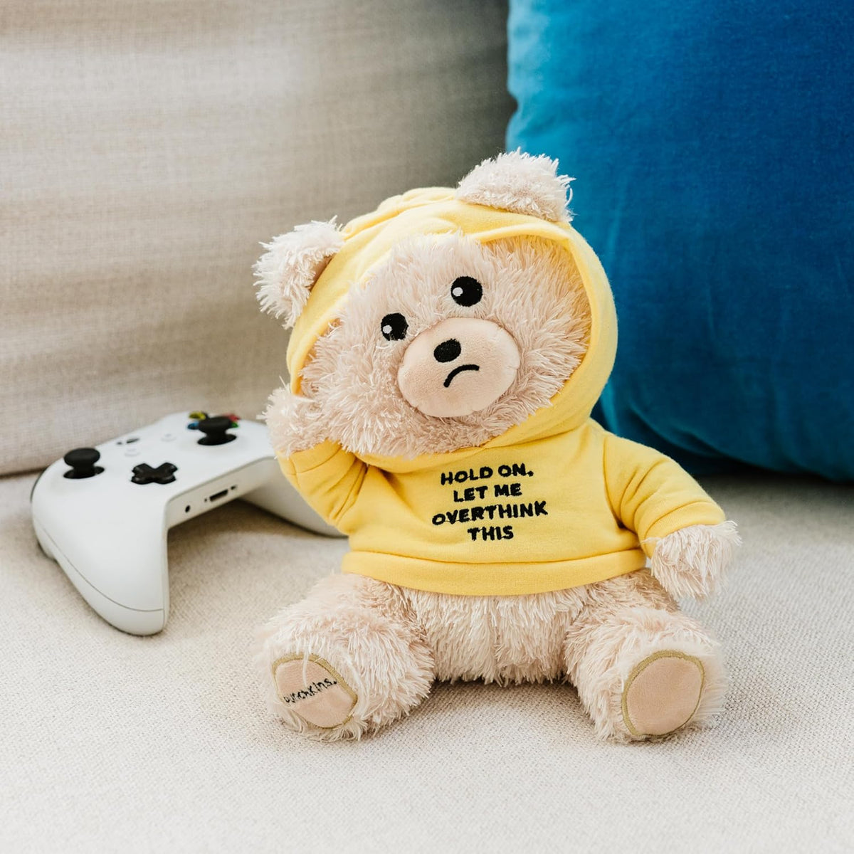 The Punchkins 🐻 "Let Me Overthink This" Teddy Bear Plushie, wearing a yellow hoodie, sits on a beige couch beside a white game controller and blue cushion—an ideal funny gift for overthinkers and any cozy space.