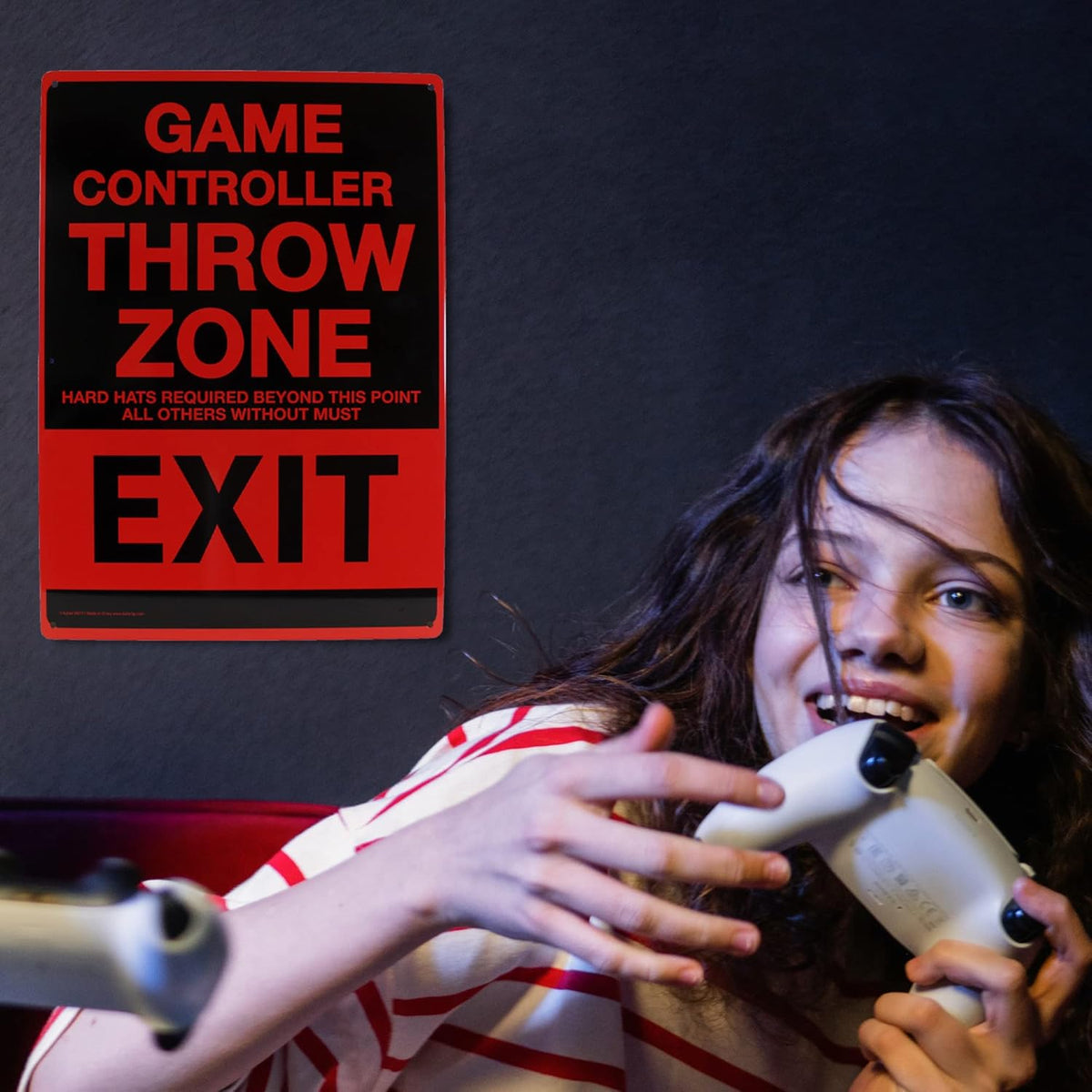 With a sly grin, the brown-haired individual holds a game controller close to their face, while in the background, the Metal Tin Sign "Game Controller THROW ZONE" Bedroom Gamer Wall Decor #F-101 is displayed.